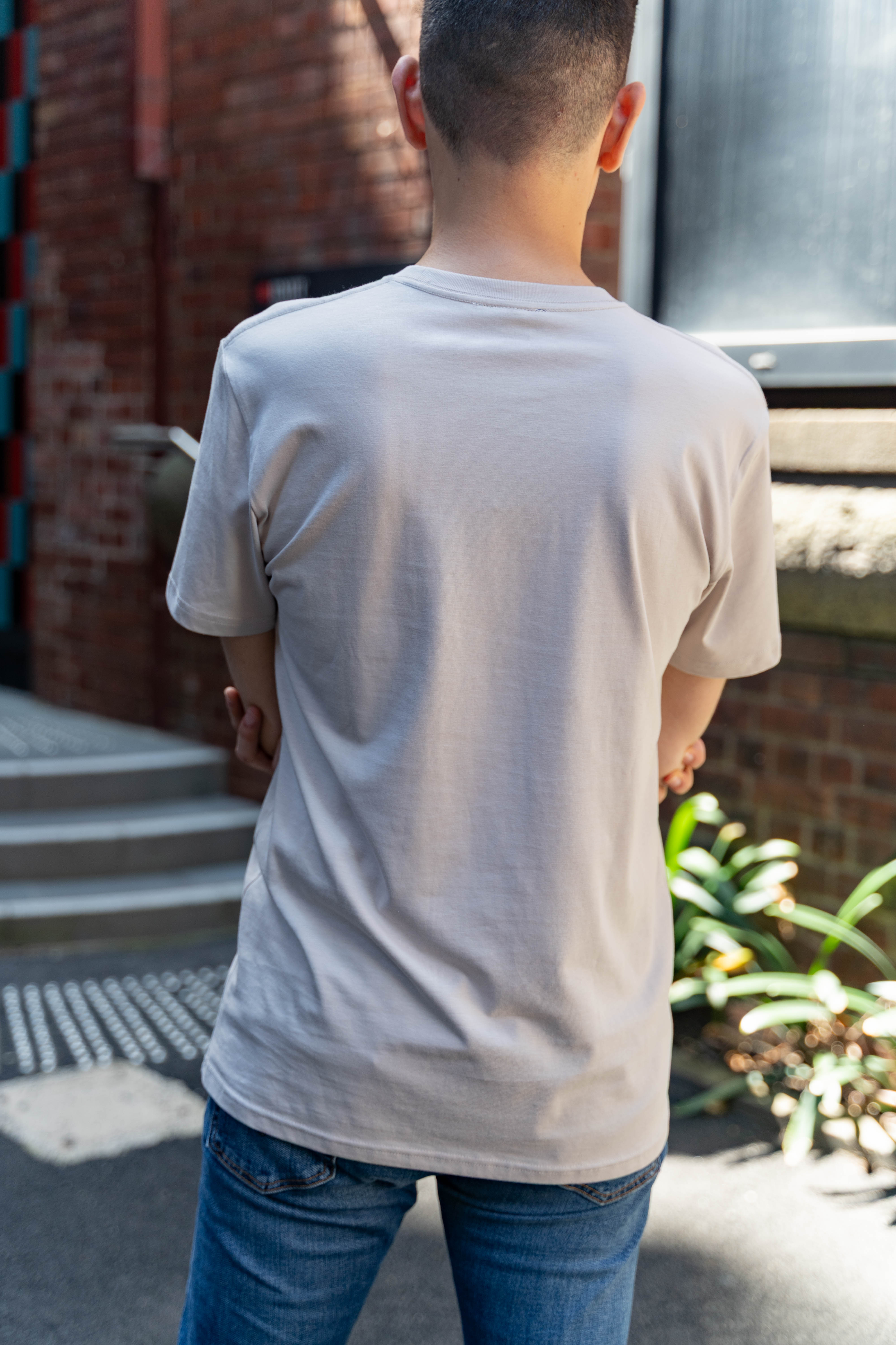 RMIT Store Pride Unity Tee in Bone, modelled by a person standing with their back to the camera against a neutral backdrop. The bone-coloured cotton t-shirt features five vibrant pride flags across the chest, visible from the rear. The tee has a relaxed fit with short sleeves and a round neckline. The model’s posture emphasises the comfortable, casual style of the tee, promoting unity and inclusivity for the LGBTIQA+ community.